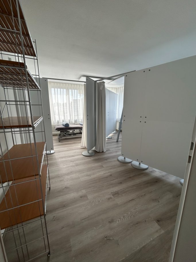 Moderner Raum mit Holzboden, Regalen und Vorhängen, helle Fenster im Hintergrund.